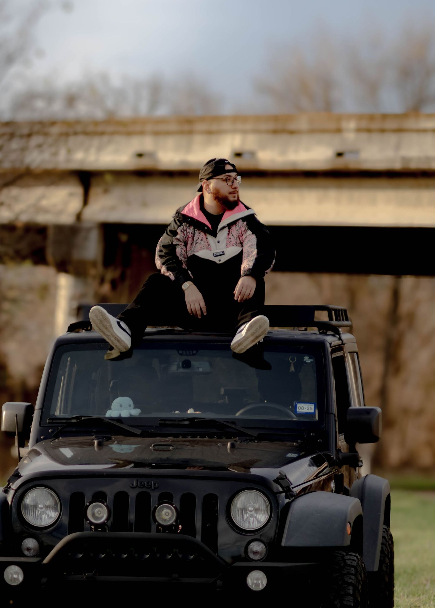 Person sitting on top of a black Jeep Wrangler wearing casual streetwear and sunglasses in an outdoor setting.