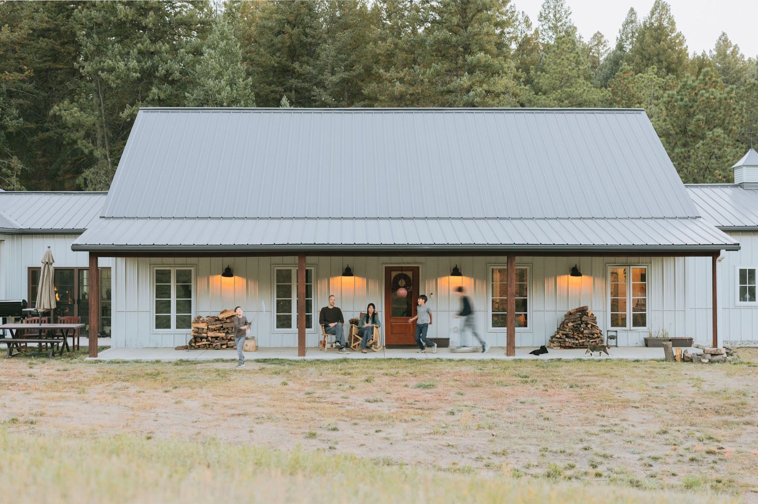 Modern farmhouse-style home with white exterior, metal roof, and welcoming front porch illuminated at dusk in wooded setting.