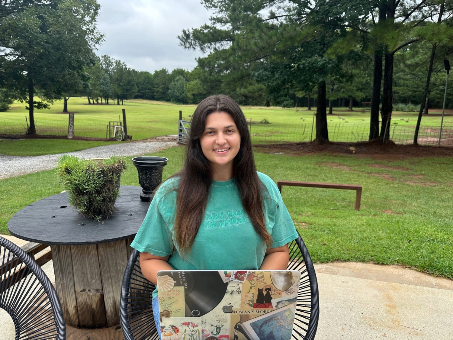 Anna Rich is an employee at Sanctuaire Atelier venue. She is a woman sitting on a patio with her laptop in front of a field of geese.