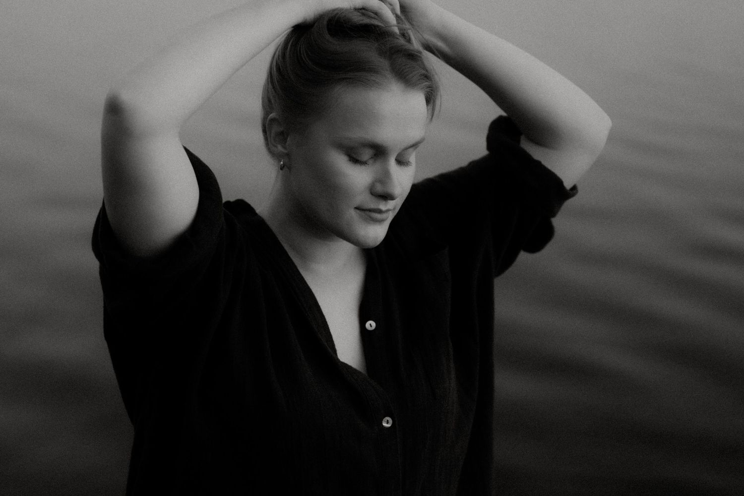 A person with long blonde hair in a black jacket stands joyfully with arms outstretched near calm water at dusk.