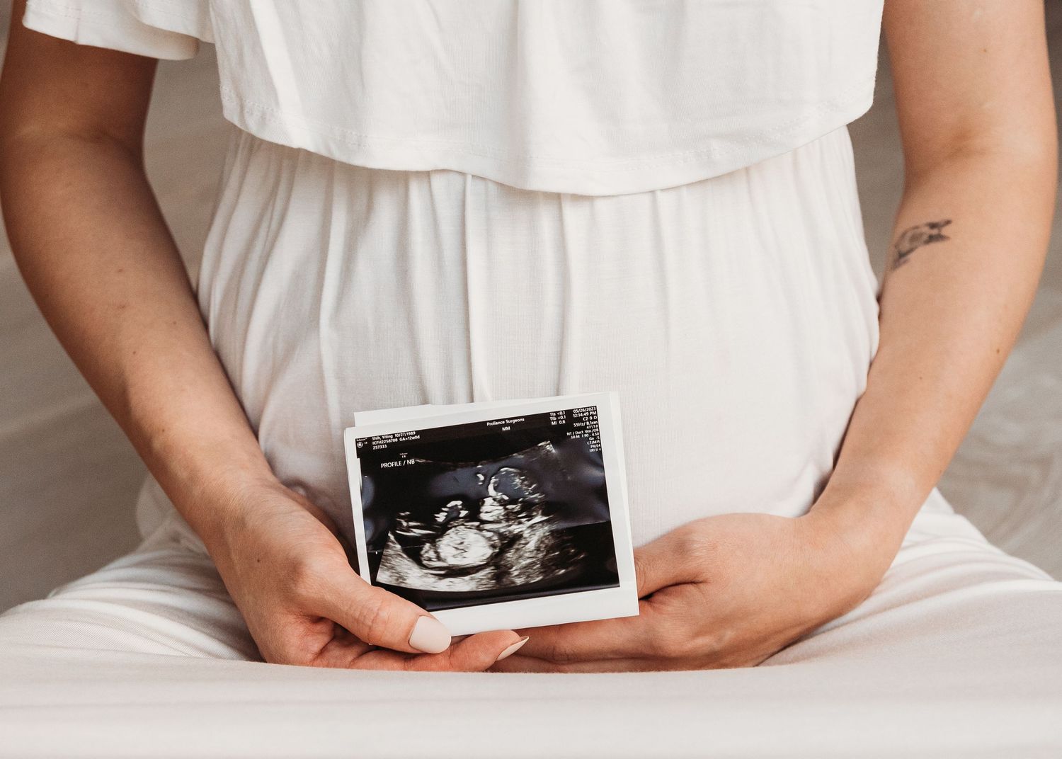 A person in a white dress holds a black and white ultrasound photo against their stomach.