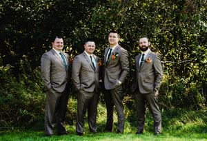 Four groomsmen in matching gray suits with boutonnieres pose together on green grass with trees in background.