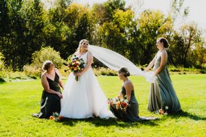 Wedding couple sits together on grassy lawn while bride's veil flows dramatically in the wind on sunny afternoon.