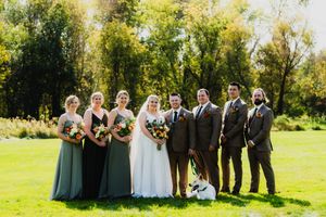 Wedding party in green dresses and dark suits poses together on sunny lawn with small dog in front.