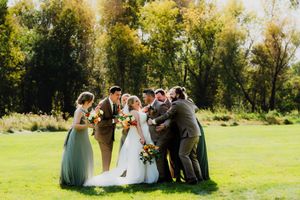 Wedding party posing for photos outdoors in a sunny green field with trees in the background.