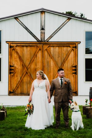 A wedding couple stands with their white dog in front of rustic wooden barn doors at a modern farmhouse venue.