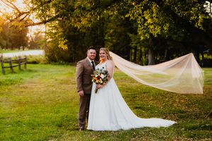 A romantic wedding couple poses in a grassy field at sunset while a long white veil flows in the breeze.