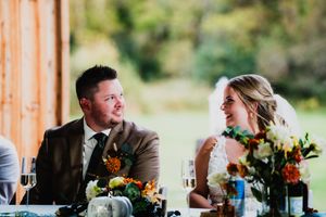 A couple shares a candid moment while seated at their rustic wedding reception table with autumn floral arrangements.