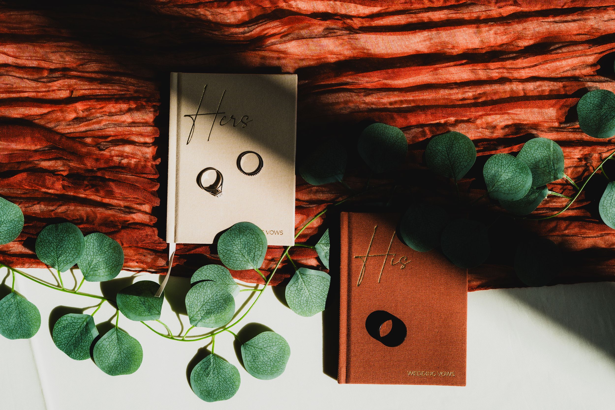 Minimalist wedding rings photographed against brown leather with green eucalyptus leaves creating an artistic arrangement.