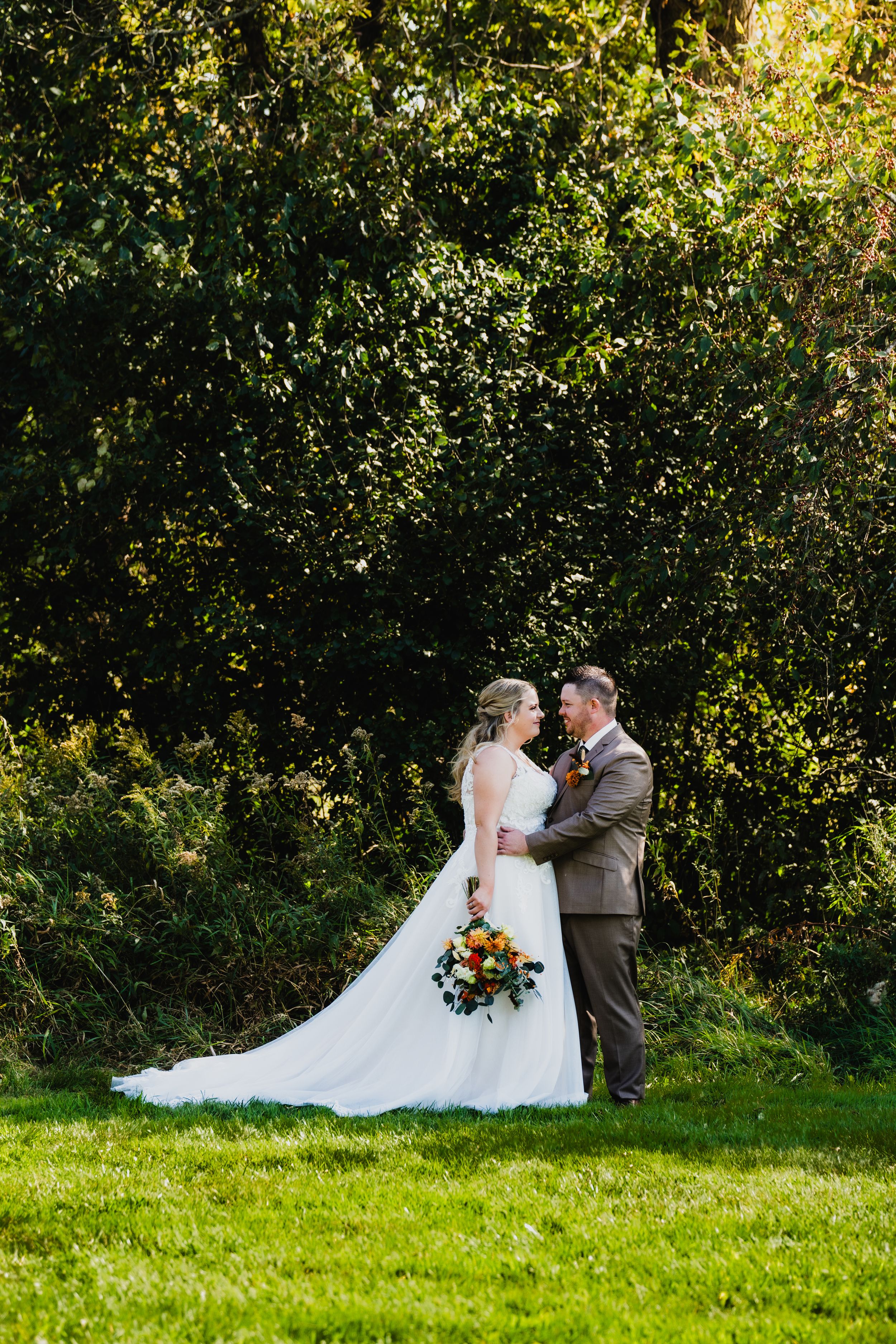 Newlywed couple embraces in a scenic garden setting with lush green grass and sunlit foliage in the background.
