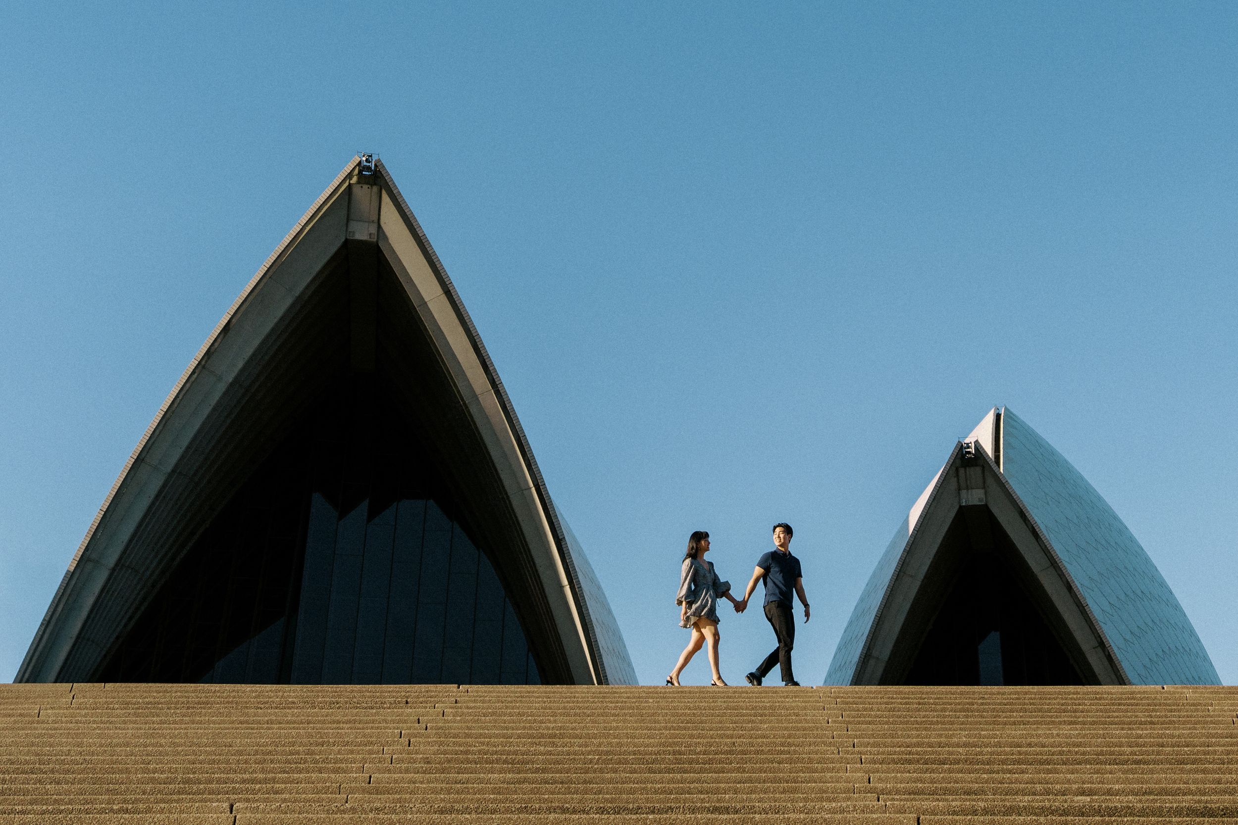 Two silhouettes walk across the iconic steps of the Sydney Opera House against a blue sky backdrop.