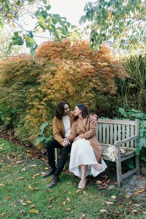 A couple sits together on a wooden bench in a fall garden surrounded by autumn foliage and changing maple leaves.