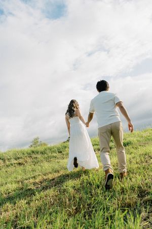 A couple walking up a grassy hillside on a cloudy day with the person in white wearing a flowing dress.