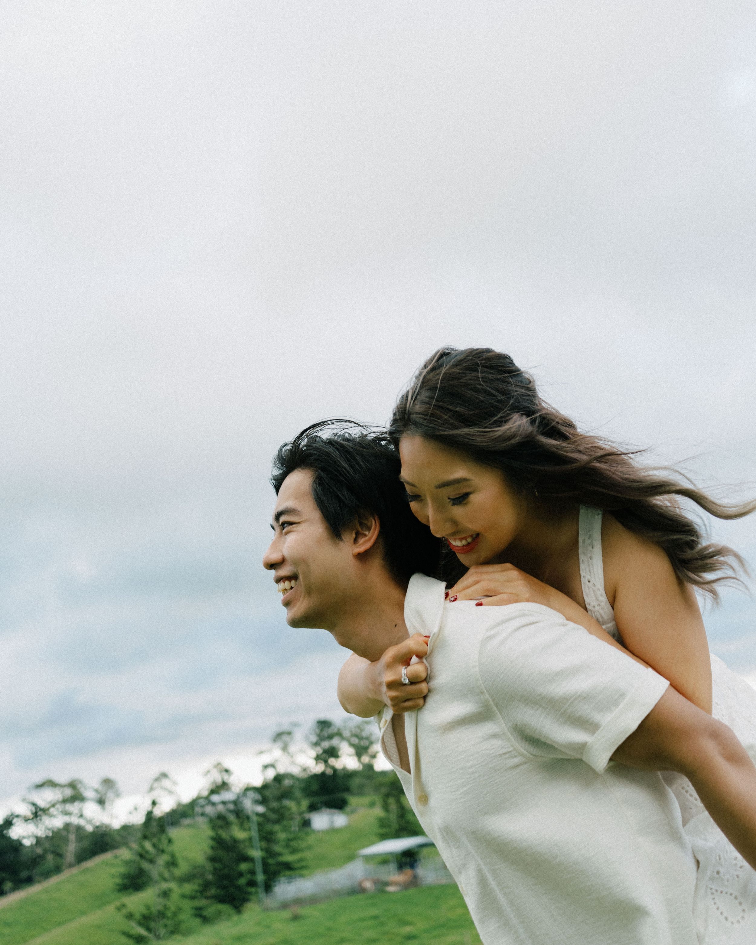 A playful couple share a piggyback ride outdoors in a green field against a cloudy sky.