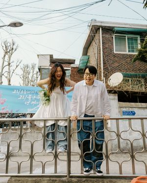 A couple in white outfits stand together behind a metal railing on a street with power lines and buildings visible.