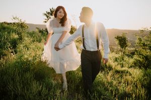 Sun-drenched couple stands in a green meadow during golden hour as soft light filters through their romantic embrace.