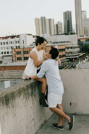 A romantic couple shares an intimate moment on a rooftop with modern city buildings in the background.