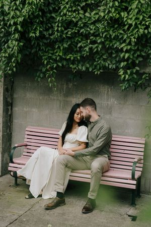 A couple shares an intimate moment while sitting together on a pink bench beneath hanging green foliage.