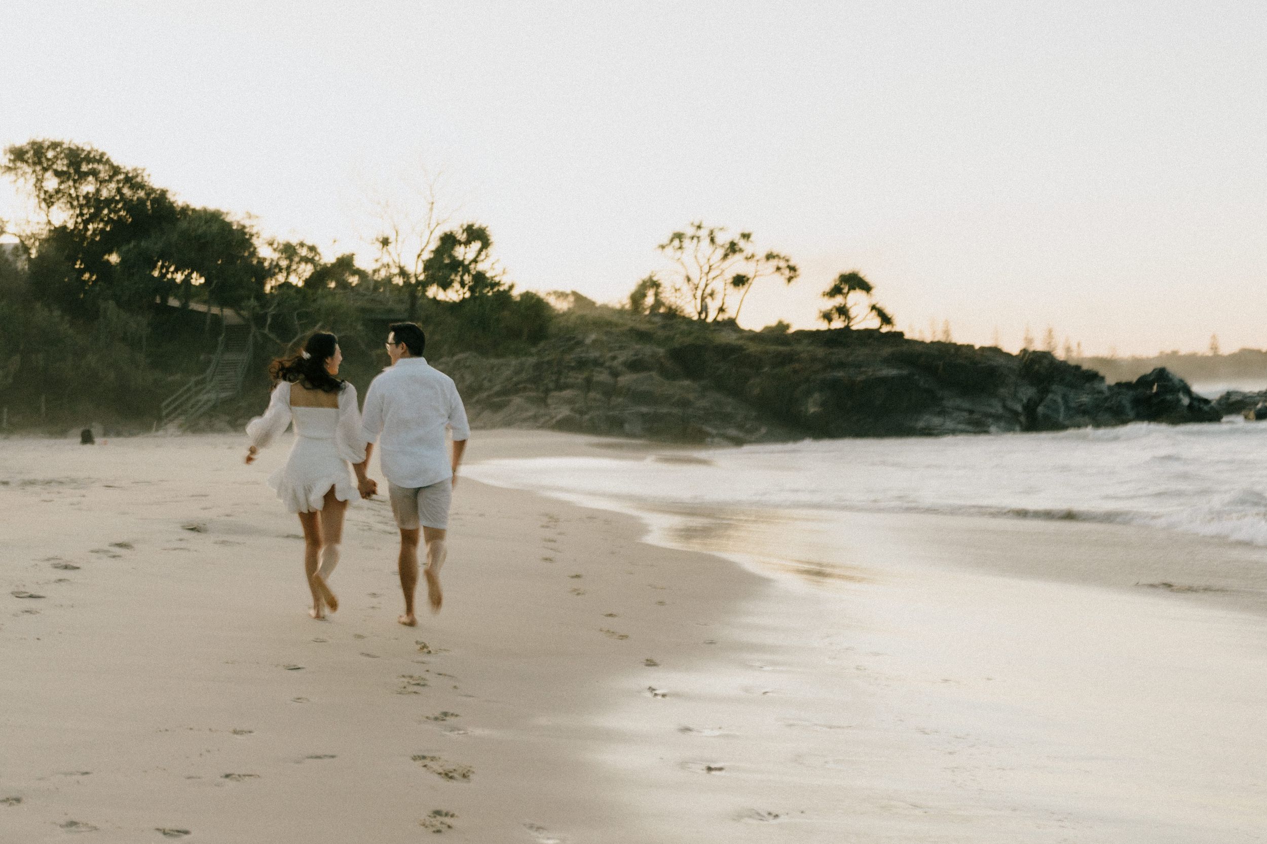 Two people walk along a scenic beach at sunset with their dog following behind on a sandy shoreline.