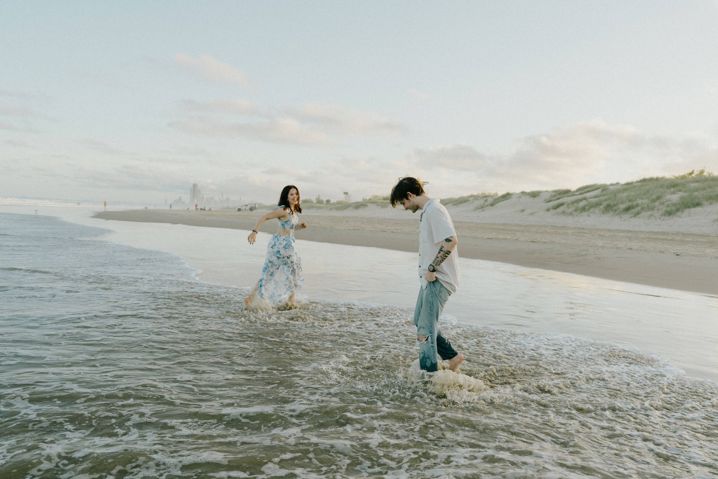Two people splash and play in shallow ocean waves along a serene beach at dusk.