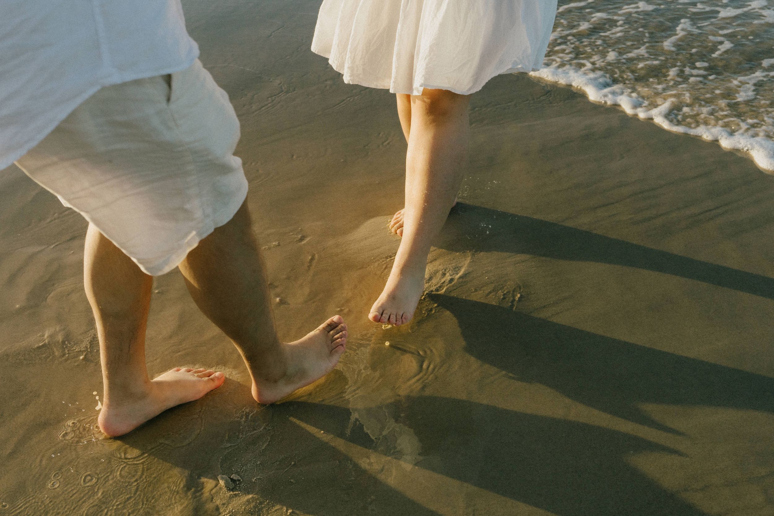Bare feet walking along a sandy beach with ocean waves and soft shadows cast in golden sunlight.