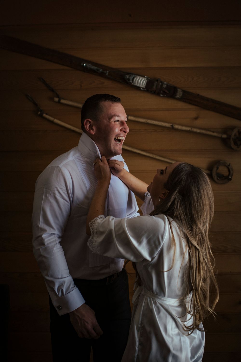 A couple in a dimly lit rustic room getting ready, with vintage tools mounted on the wooden wall behind them.