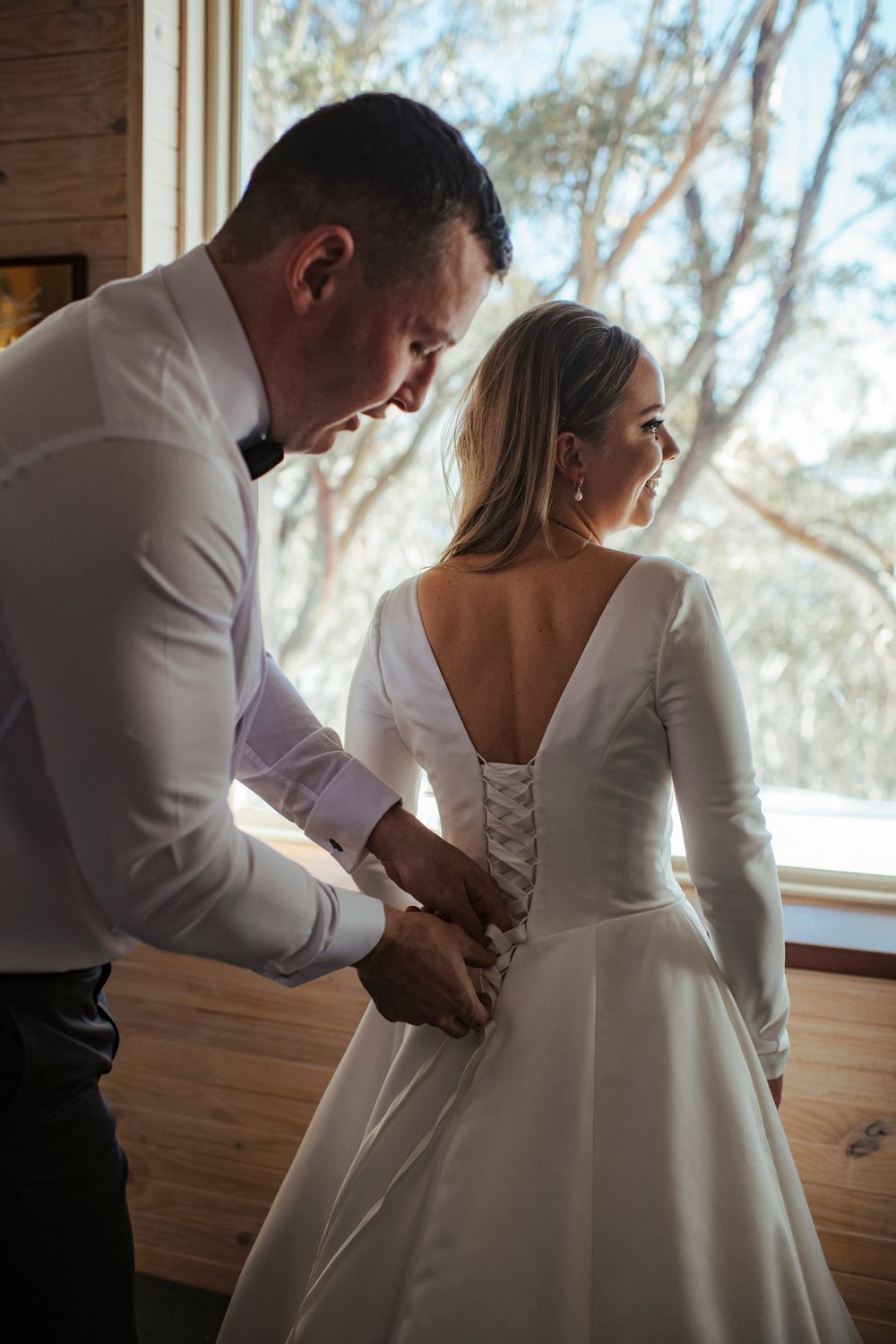 A detailed shot of someone helping to lace up a white wedding dress with long sleeves by a sunlit window.