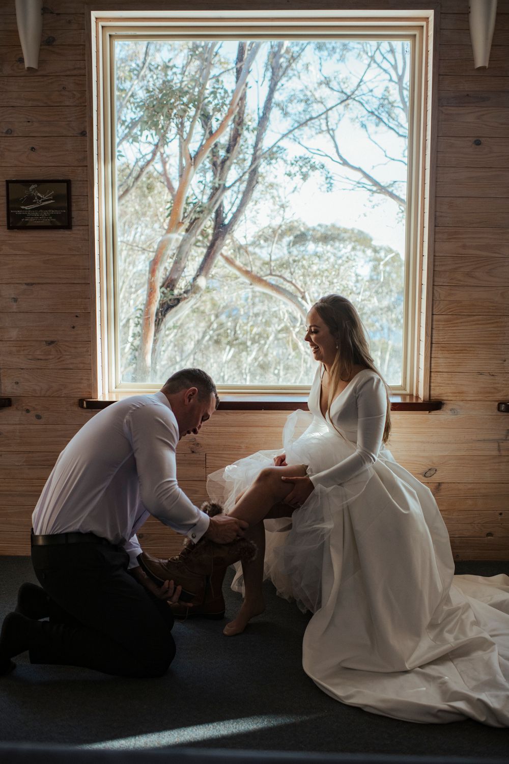 A tender moment is shared beside a large window in a rustic wooden room, with sunlight streaming through trees outside.