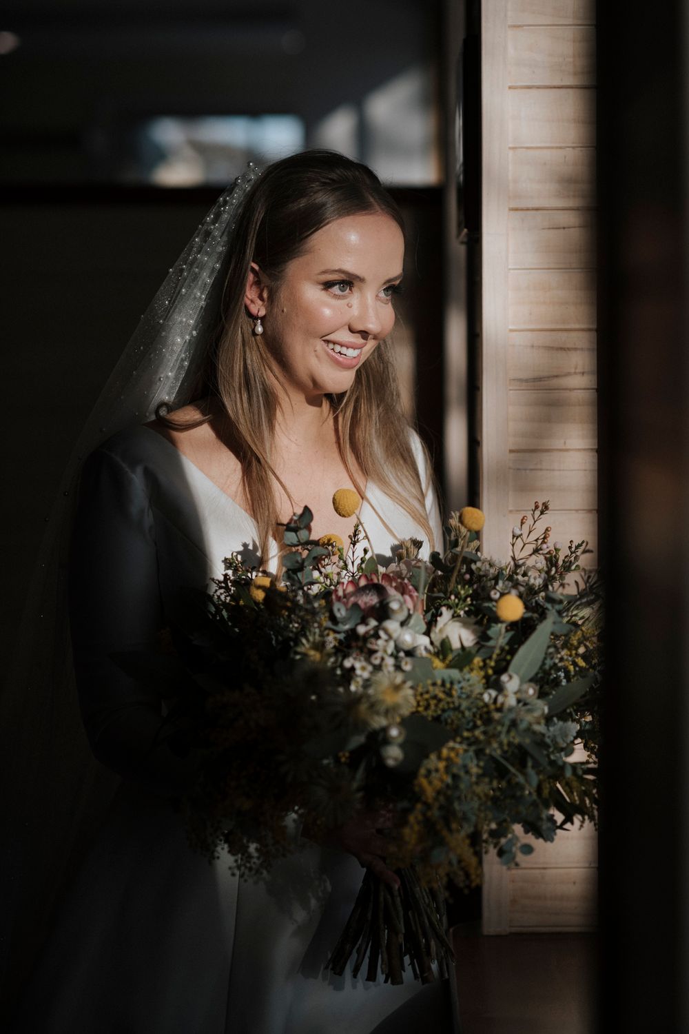 A bride in a white wedding dress holds a beautiful rustic bouquet with yellow and purple flowers beside a window.