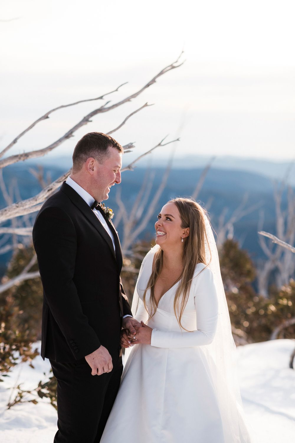 Winter wedding photos show a couple laughing together in the snow with mountains in the background.