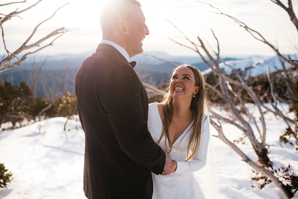 A couple shares a joyful moment in formal attire against a snowy mountain backdrop on a bright sunny day.