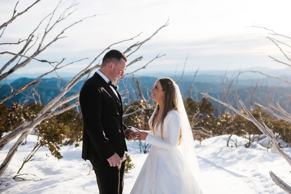 A winter wedding couple stands on a snowy mountaintop with bare trees and a scenic mountain vista in the background.