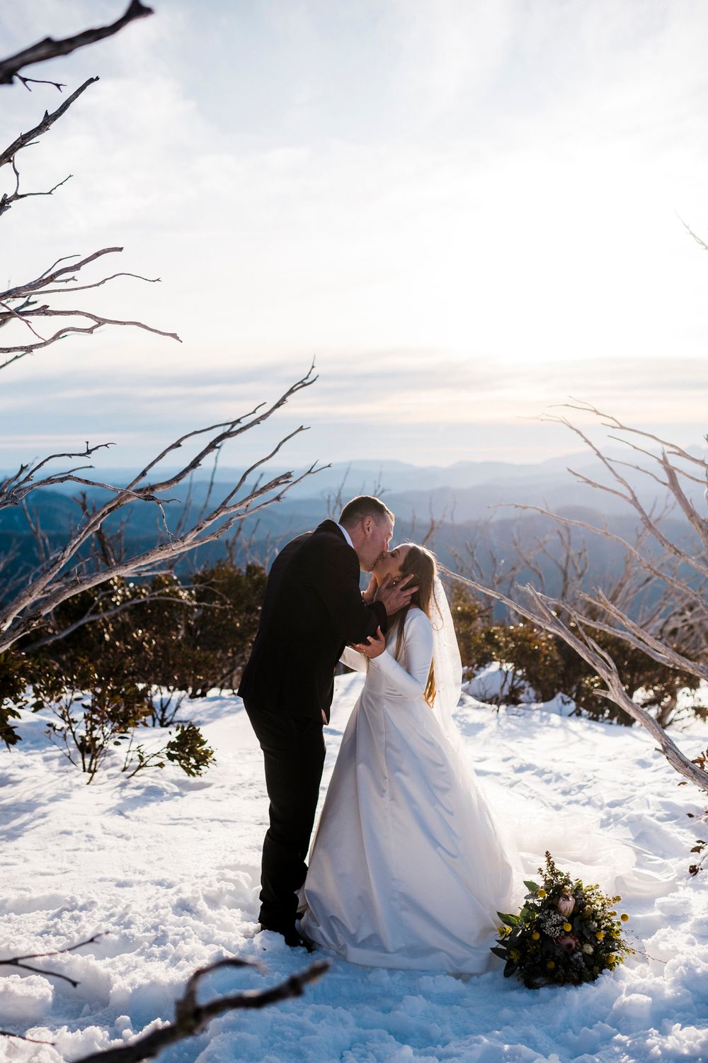 A romantic winter wedding photo showing newlyweds sharing an intimate moment in the snow-covered mountains at sunset.