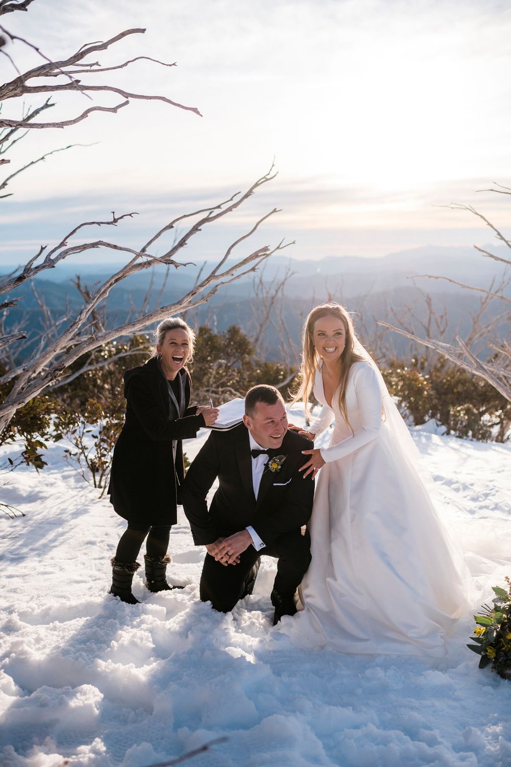 A wedding party of three poses playfully in the snow on a mountaintop with scenic winter views.