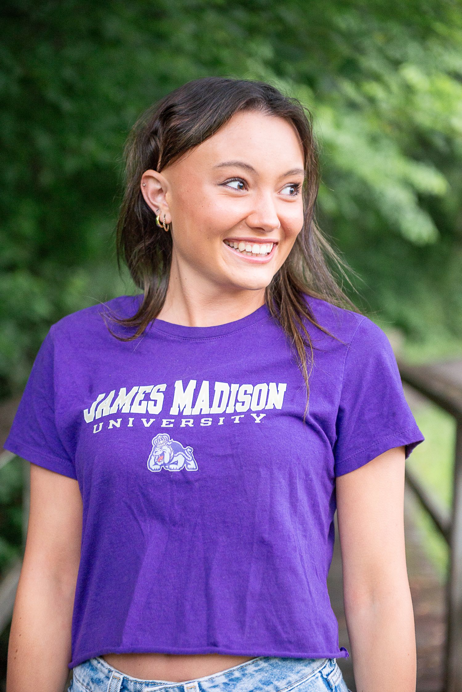 A James Madison University fan wearing a purple crop top t-shirt with the school logo smiles outdoors against green foliage.