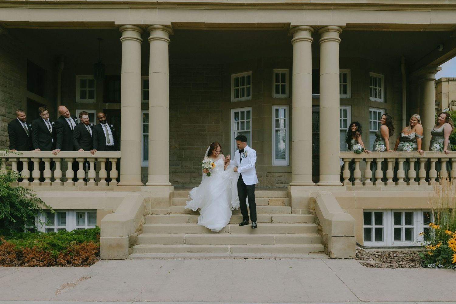 A couple stands beneath a stone colonnade in an elegant garden setting during a wedding celebration.