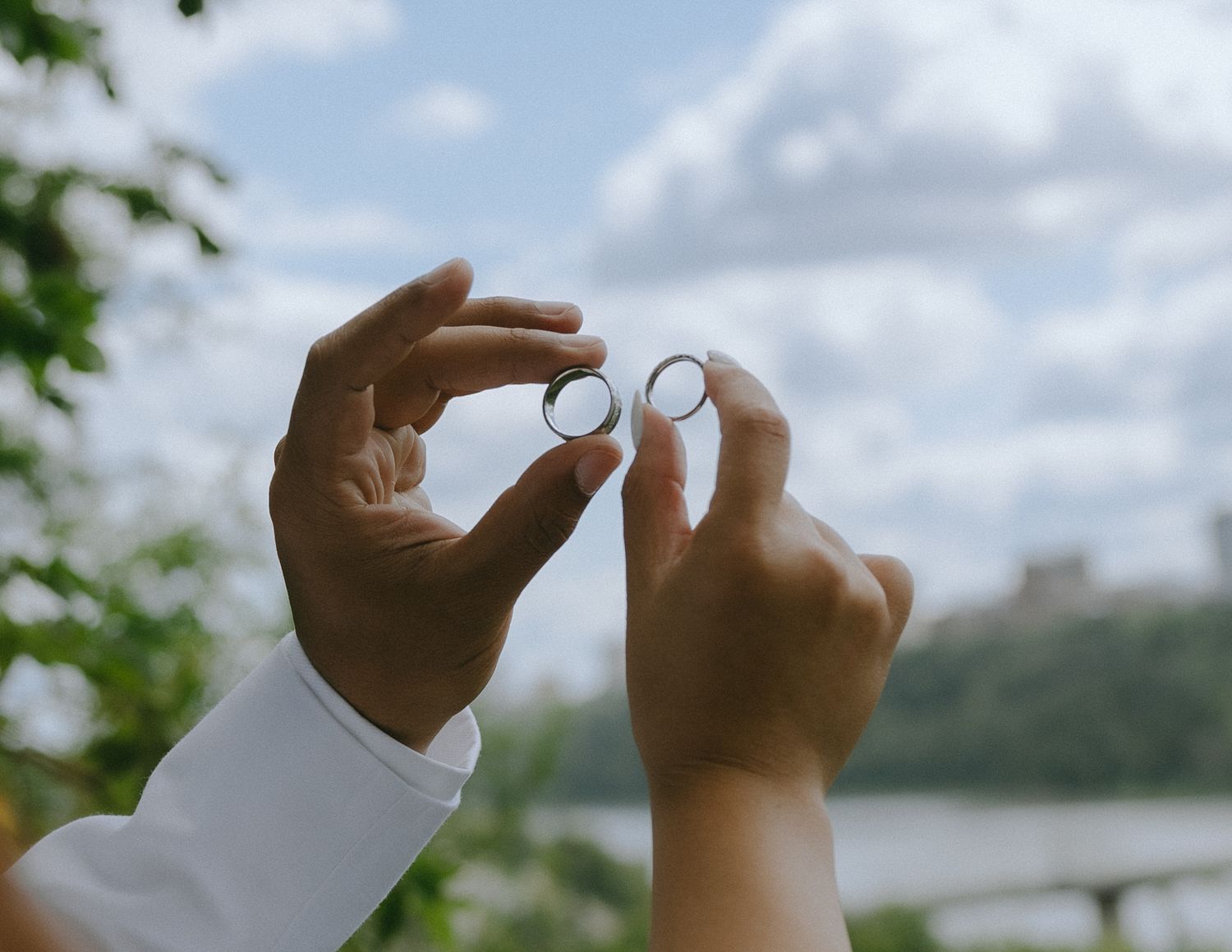 Hand holding a wedding ring against a blurred green outdoor background with soft bokeh effect.