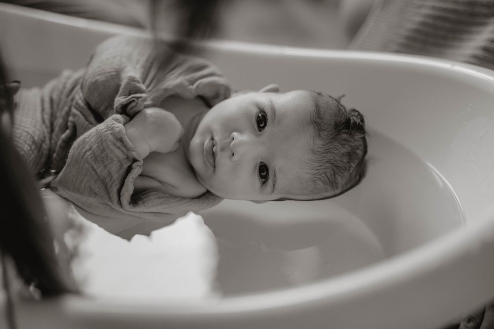 Black and white photo of a small child laying in a white bathtub, looking up at the camera with a gentle expression.