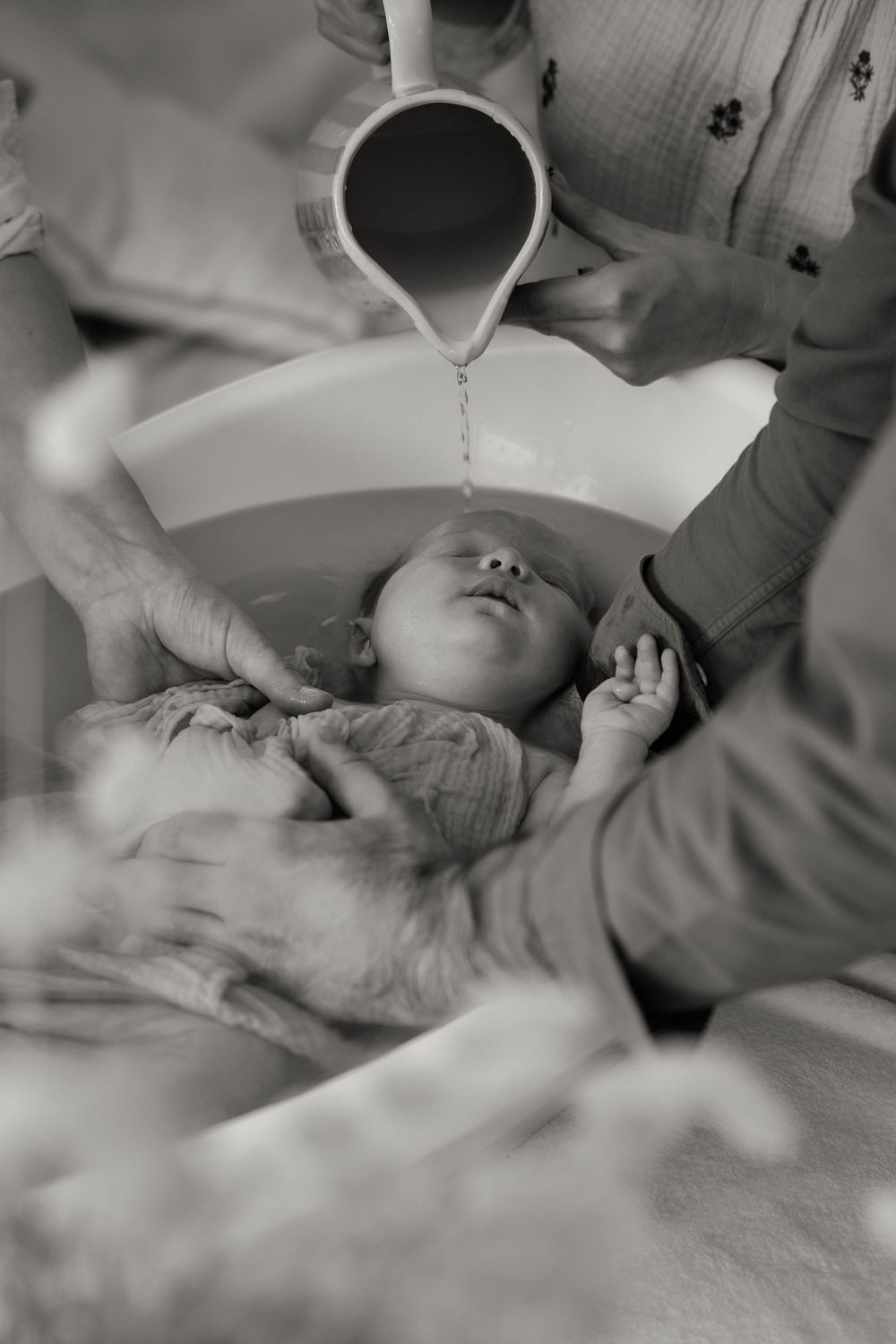 Black and white photo of a newborn baby being baptized with water poured from a white pitcher.