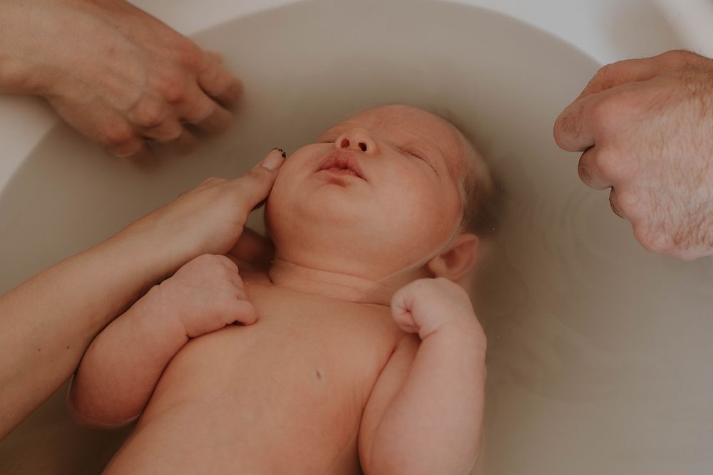 Newborn baby being gently bathed in a white tub, supported by caring adult hands during first bath.