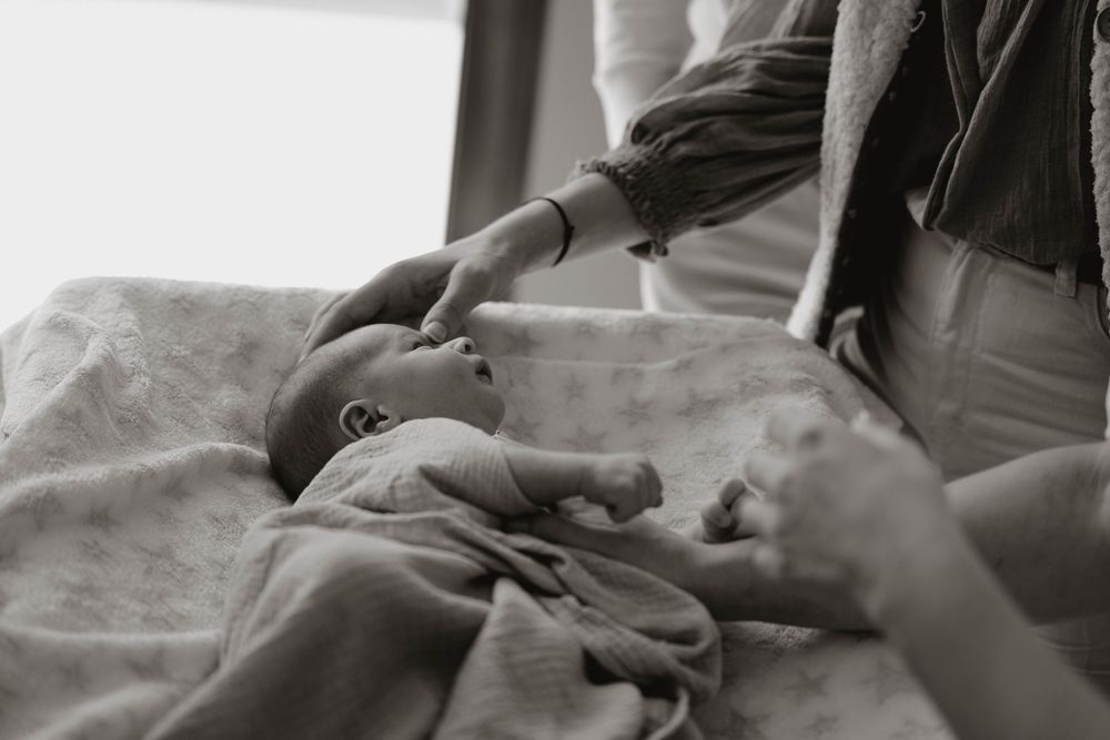 Black and white photo of a newborn baby lying on a blanket while a parent gently touches its face.