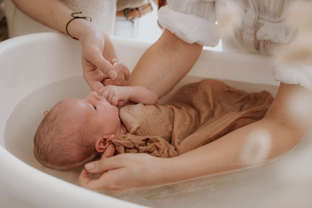 Newborn baby wrapped in brown muslin cloth being gently bathed in a white baby bathtub by caring hands.