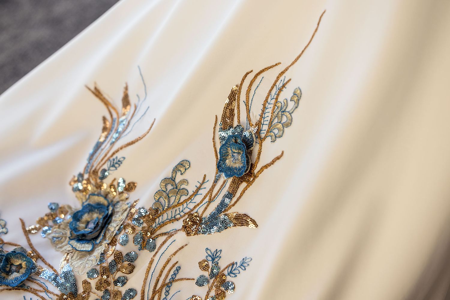 Elegant beaded embroidery detail featuring blue gemstones and golden thread work on ivory fabric.