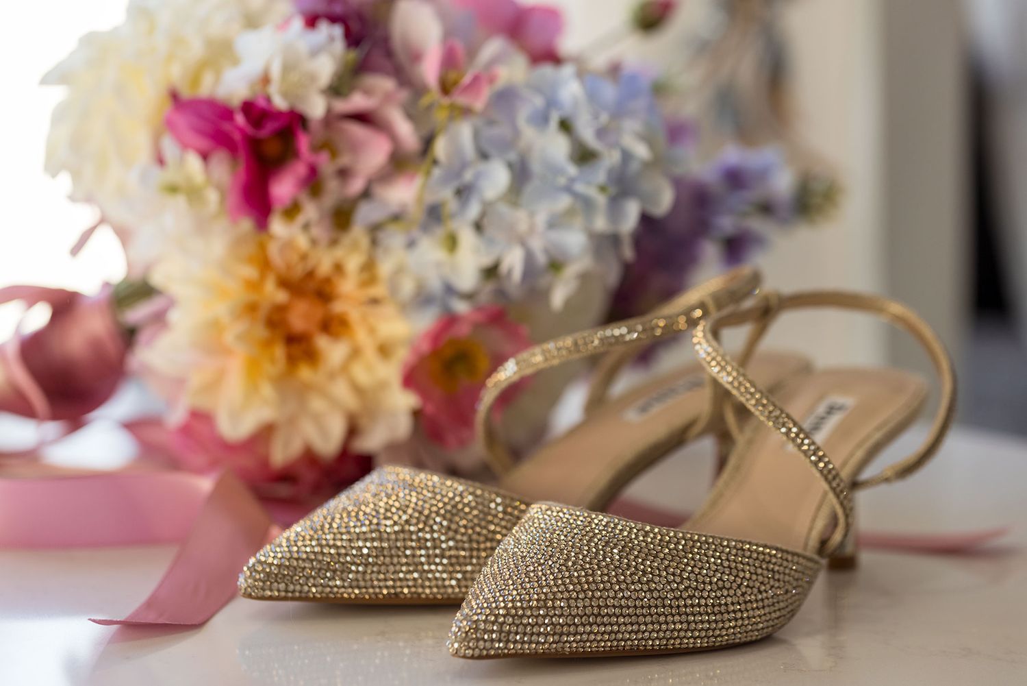 Sparkly gold wedding shoes with ankle straps sit beside a colorful bouquet of flowers and pink ribbon.