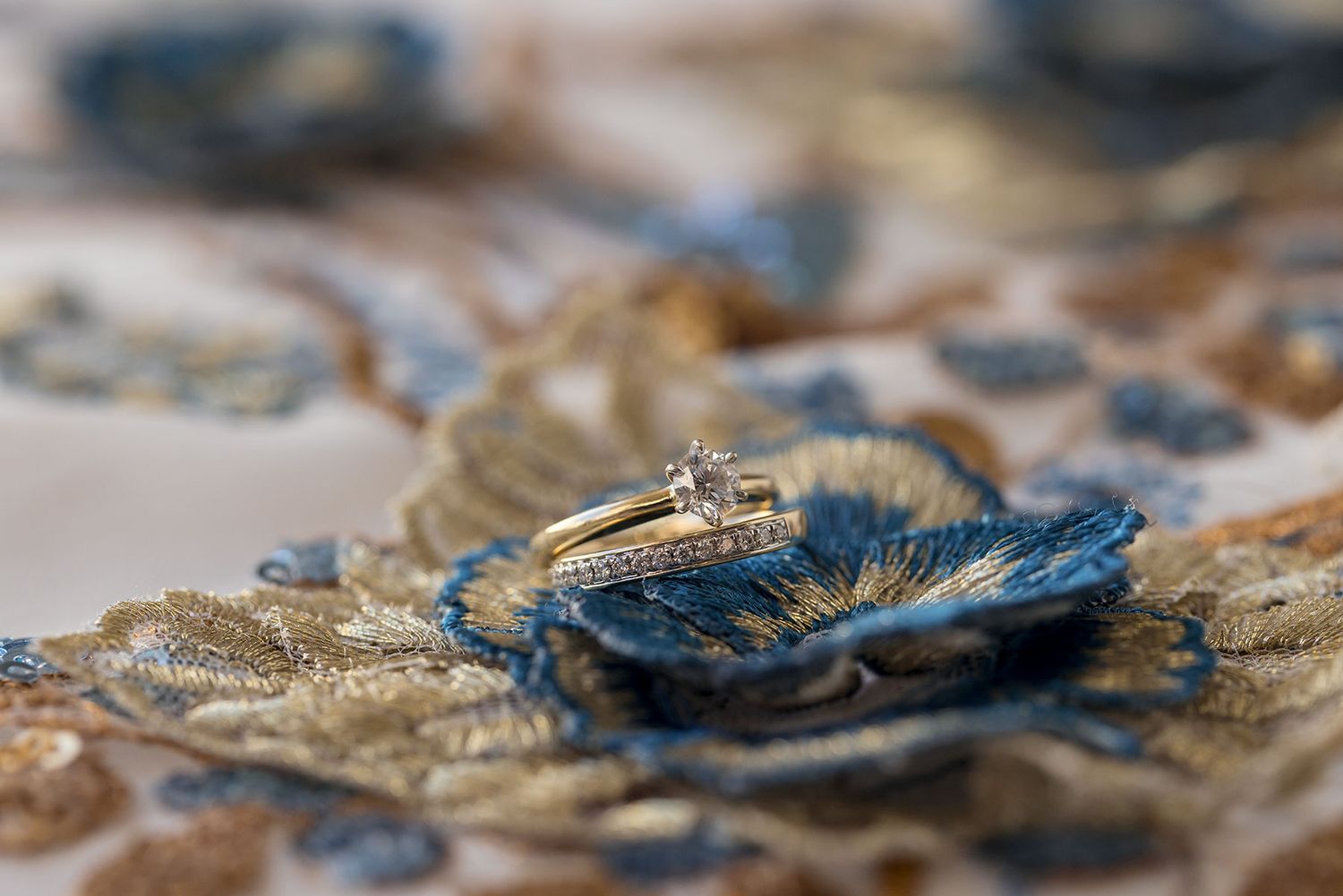 Wedding rings rest on intricate blue and gold floral embroidered fabric on a Brides East meets Dress Gown with shallow depth of field.