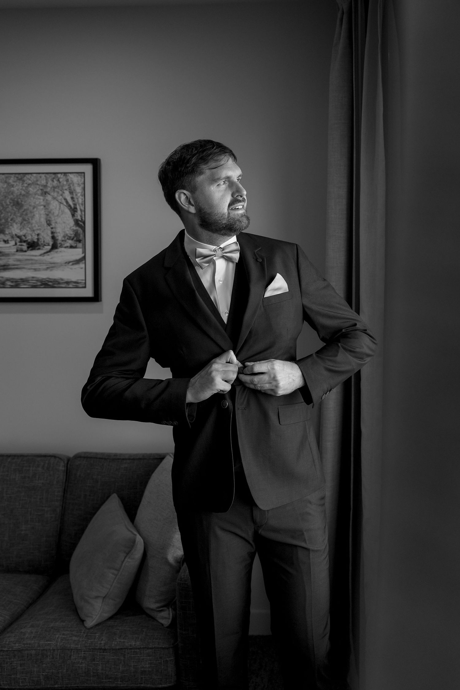 Black and white portrait of groom in formal tuxedo and bowtie adjusting his jacket while looking out window at Quest Albury.