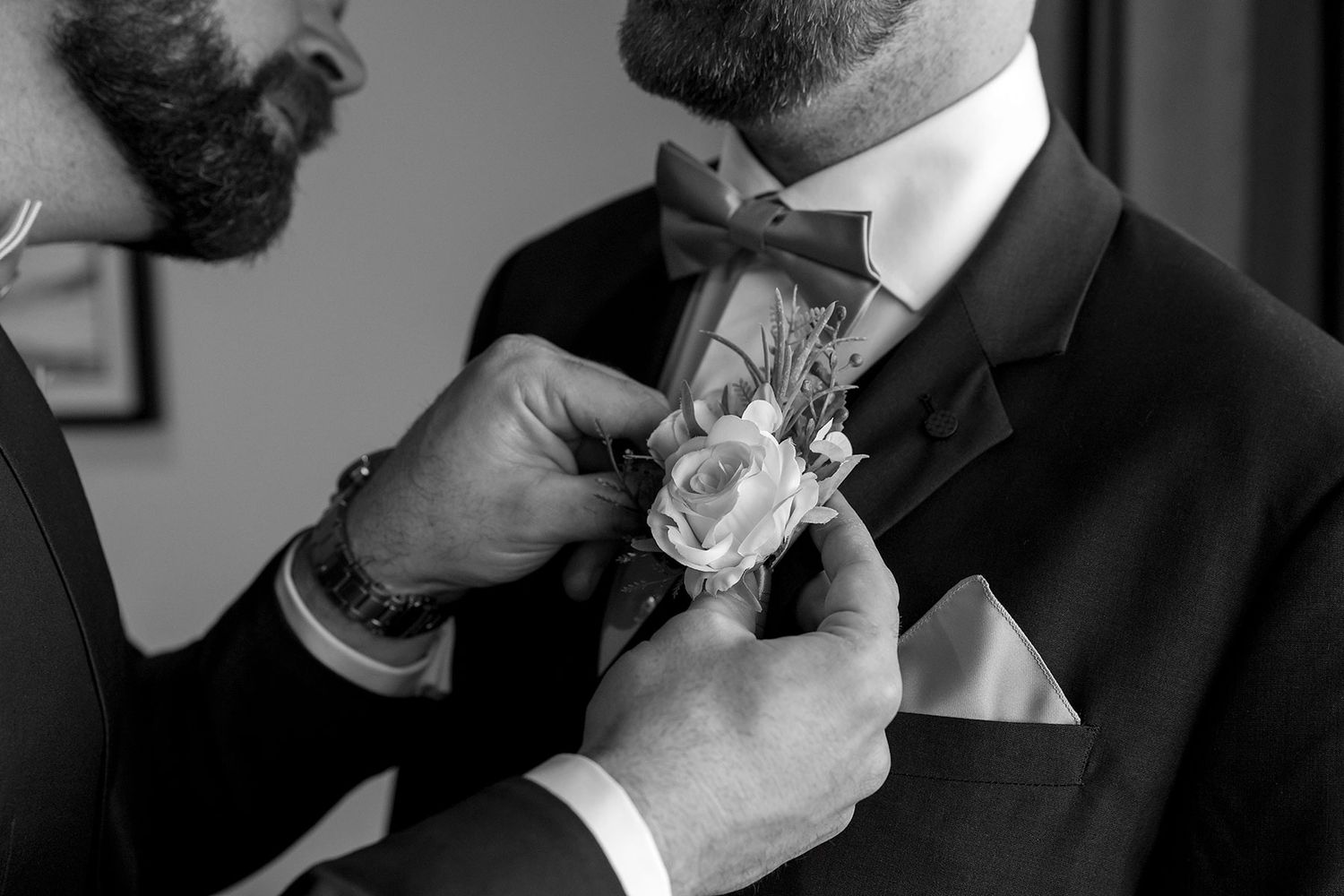 The Best man helps adjust a white rose boutonniere on a groom's lapel in this black and white wedding photo at Quest Albury.