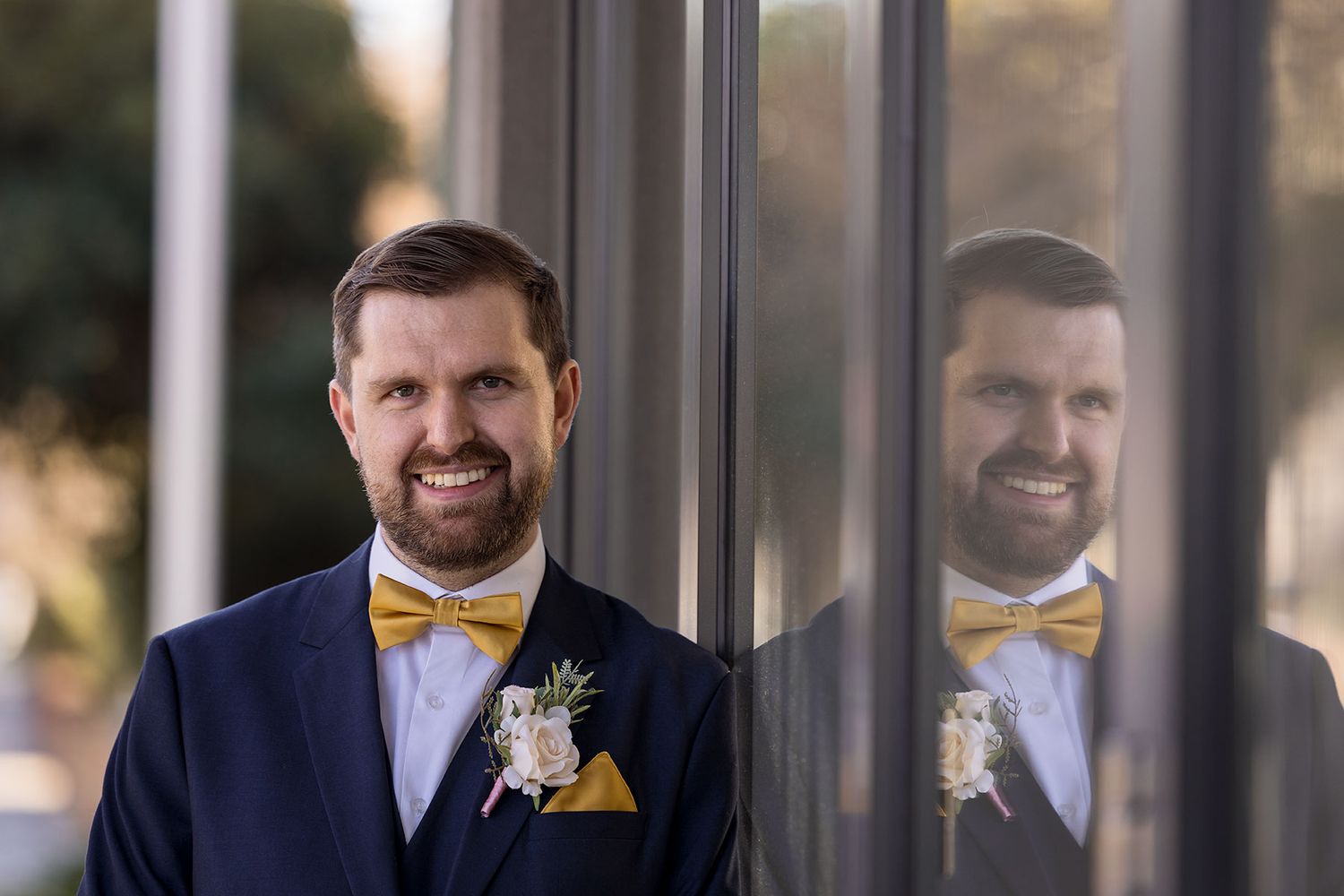 A groom in a navy suit with yellow bowtie and boutonniere stands next to a reflective window surface at Quest Albury.