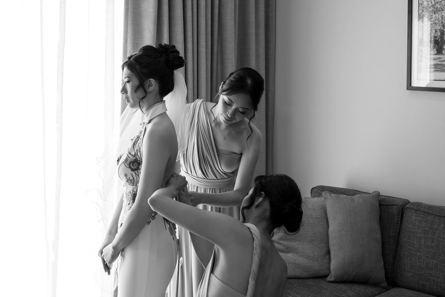 Bridesmaids help a Bride with the back of her 'East meets Dress' wedding gown at in a bright hotel room at Quest Albury.