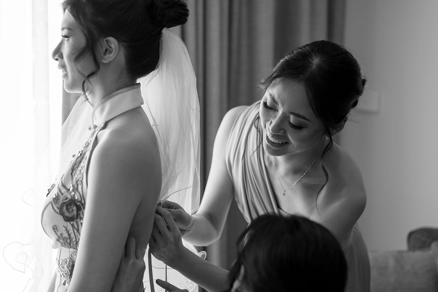 Bridesmaids help a Bride with the back of her 'East meets Dress' wedding gown at in a bright hotel room at Quest Albury.